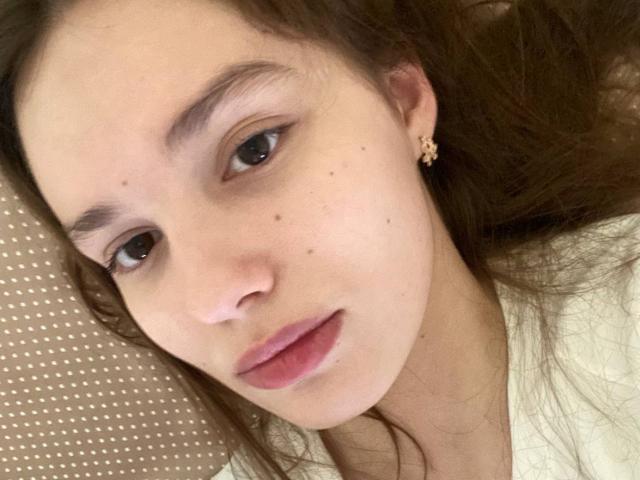 HanaArt's face, showing brown eyes, pink lips, and small moles on her cheek and nose, with brown hair and a small gold earring, lying on a beige pillow.