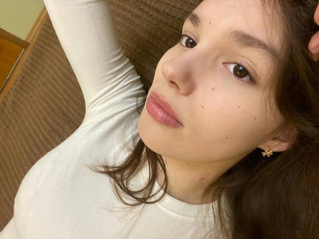 HanaArt has light skin, brown eyes, and pink lips, wearing a white top, with brown hair and small earrings, showing a relaxed, slightly smug face in a selfie.