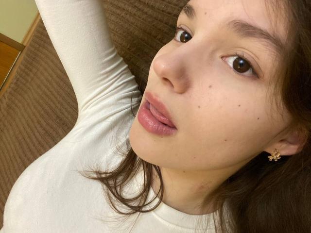 HanaArt with her pouty, shiny pink lips, small moles on her face, brown eyes, and white top, showing a slightly raised arm. Brown hair, small flower earring.