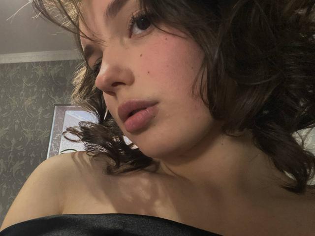 HanaArt's face, showing a slight pout with parted lips, curly brown hair, and freckles. She's wearing a black strap, and her skin is fair.