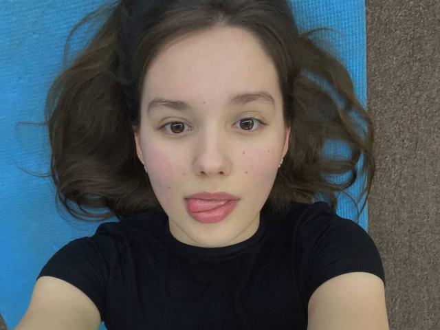 HanaArt with puffy pink lips, lying on a blue mat, wearing a black shirt, brown hair splayed, brown eyes, light skin, small nose, and subtle freckles.