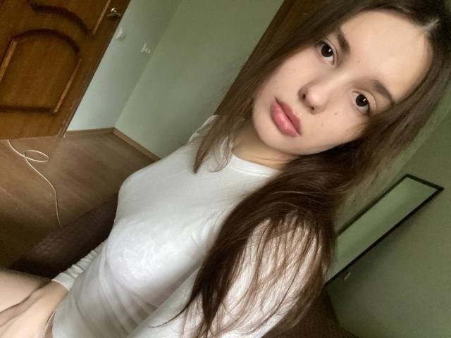 HanaArt with long brown hair, wearing a white shirt, showing small tits, sitting, looking at the camera, in a dimly lit room, with a wooden door in the background.