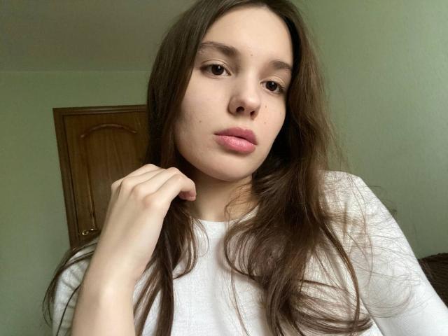 HanaArt has long brown hair, pink lips, and light skin, wearing a white shirt, with her hand near her chin, looking slightly bored. Wooden door in background.