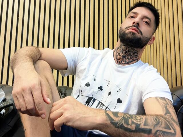 TommyDum with a beard, tattoos on neck and arm, white shirt, sitting with knee up, looking at camera, wooden vertical background, short black hair, relaxed, confident, sexy.