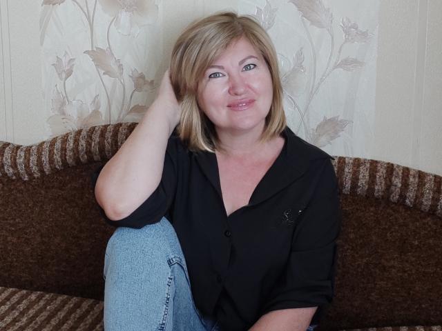 Leonie, mit blonden Haaren, blauen Augen und einem leichten Lächeln, trägt ein schwarzes Hemd und blaue Jeans und sitzt auf einem braunen, gestreiften Sofa, wobei sie eine Hand in ihrem Haar hat..