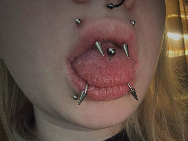 EmmaKoven's face with multiple piercings: silver studs in upper lip, lower lip, and tongue, and sharp metal spikes in bottom lip. Tongue is out. She's got blonde hair.