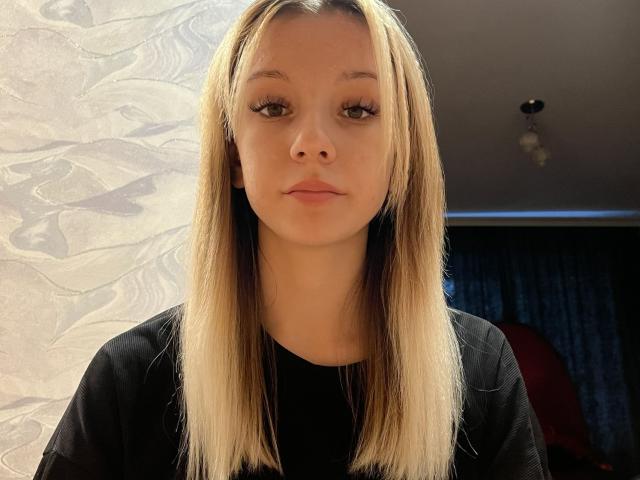 CherelleBettle tiene el cabello rubio y lacio, piel clara y labios carnosos con un maquillaje sutil. Lleva una camisa negra y mira directamente a la cámara con una expresión neutra..