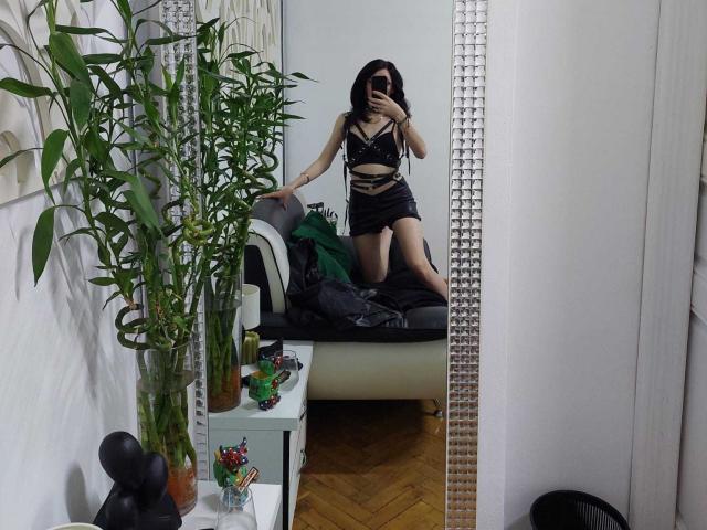 VanessaDomy kniet auf einer schwarzen Ledersofa, trägt ein schwarzes Spitzenoberteil und schwarze Shorts, wobei ihr Hintern zu sehen ist. Ein Spiegel spiegelt sie wider. Im Hintergrund stehen Bambuspflanzen in Töpfen und eine weiße Wand..