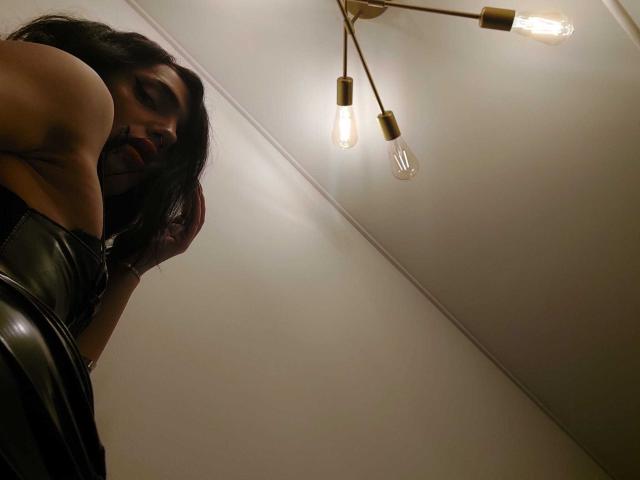 VanessaDomy, com cabelo escuro, olhando para baixo, vestindo calças de couro pretas, sob uma lâmpada de teto com lâmpadas aparentes, com suas nádegas e costas visíveis, em um quarto com pouca luz..