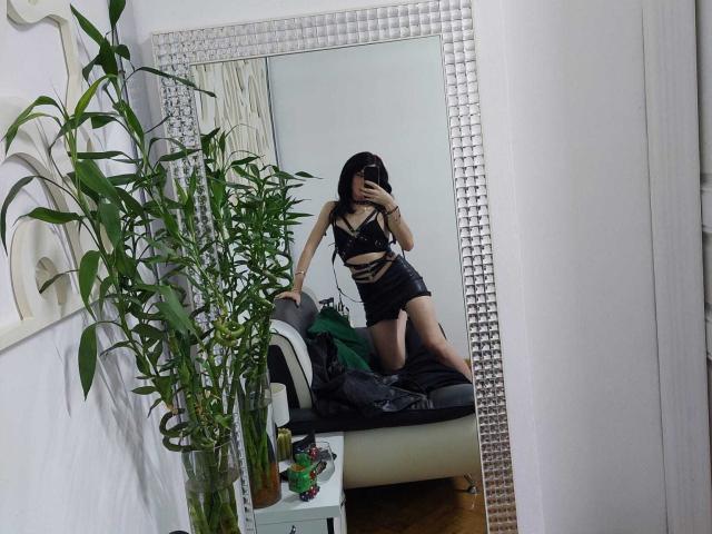 VanessaDomy, com cabelo preto, seios pequenos e um traseiro esguio, vestindo uma blusa e shorts de couro preto, ajoelhada em uma cama, refletida em um espelho texturizado, segurando um telefone..