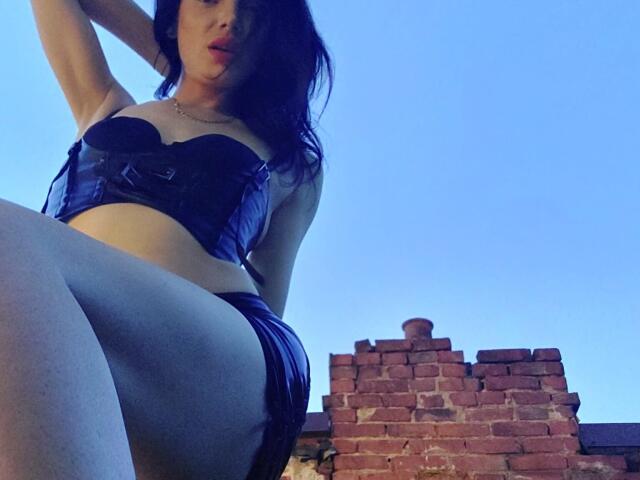 VanessaDomy with big tits, wearing a black strapless corset and black panties, sitting on brick ruins, legs spread, blue sky background, looking down with dark wavy hair.
