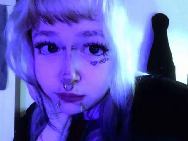 EmmaKoven with blue-lit face, pink and white hair, nose ring, lip ring, and heart tattoo under right eye, looking directly at camera. Her ass is in the background.