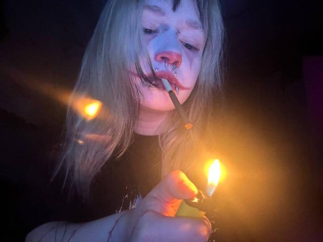 EmmaKoven with blonde hair, nose piercings, and a black shirt, smoking a cigarette with a lit match in her hand, glowing matchlight in dark, moody lighting.