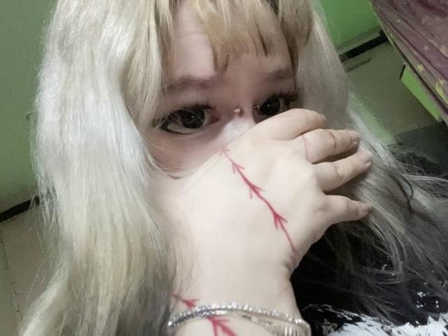 EmmaKoven with pale blonde hair, black-tipped, covering her mouth with a hand showing red scratch marks. She has a nose ring and a silver bracelet on her wrist.