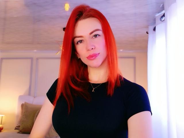 Leahred has bright red hair, a cute smile, and large tits in a black shirt, wearing a necklace, with pink lipstick, standing in a well-lit room.