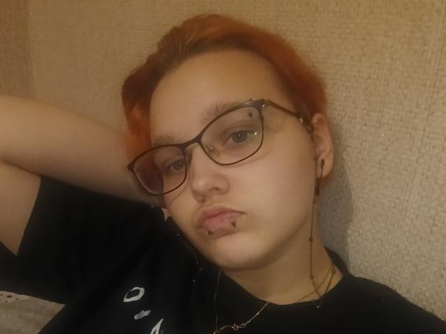 FidelaSpacek with red hair, glasses, nose piercings, and lip piercing, wearing a black shirt, has a pouty face, and is resting her arm behind her head.