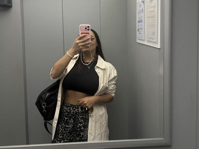 GalaMalkova with long brown hair, wearing a white shirt open to black crop top, showing her tits, and black/white patterned pants, holding a pink phone in a mirror.
