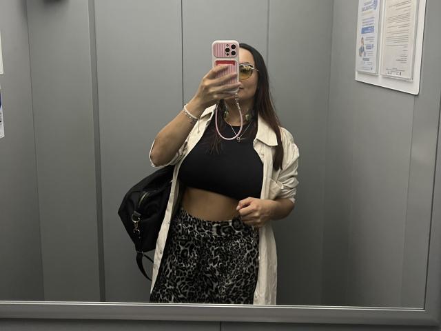 GalaMalkova with brown hair, wearing sunglasses, white coat, black crop top, and leopard print pants, holding pink phone, black bag, in mirror selfie, showing her tits and belly.