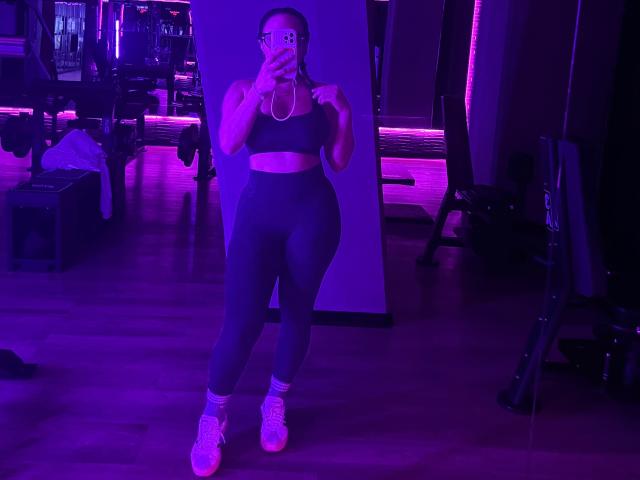 GalaMalkova in a gym, wearing a black sports bra, high-waisted gray leggings, white socks, and pink sneakers, holding a phone, with purple lighting.