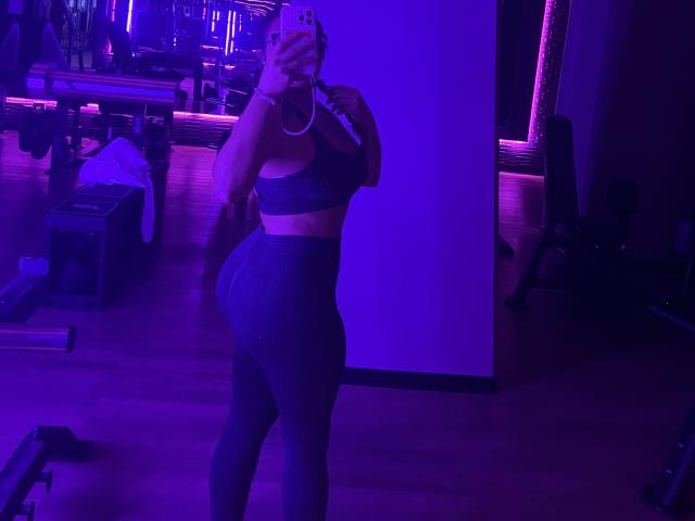 GalaMalkova with curvy, thick-ass in gray sports bra and high-waisted leggings, taking a mirror selfie in purple-lit gym, holding phone with pink case.