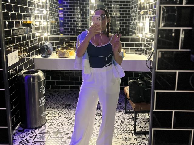 GalaMalkova stands in a black-tiled bathroom, wearing a navy top, white pants, and a white jacket, holding a phone to her face, with braids and a pink earbud.