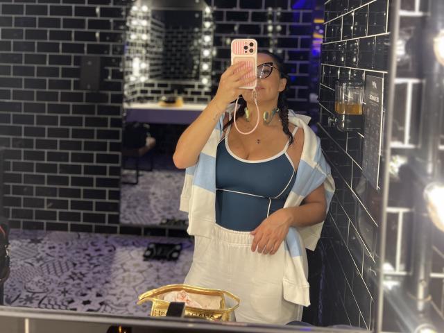 GalaMalkova with titties in a blue sports bra, white shorts, and a light blue and white towel, taking a mirror selfie, wearing glasses and a necklace, in a bathroom.