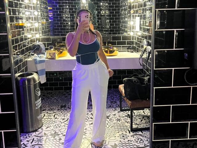 GalaMalkova with tits in a dark green tank top, white high-waisted pants, holding phone in mirror selfie, black tiled walls, black and white patterned floor.