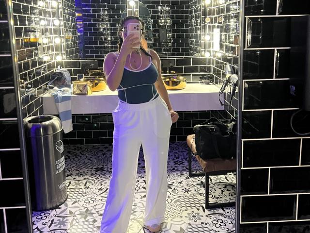 GalaMalkova with big boobs, wearing a black top, white high-waisted pants, holding phone, standing in black-tiled bathroom, mirror selfie, dark skin, wet hair, wearing a ring.