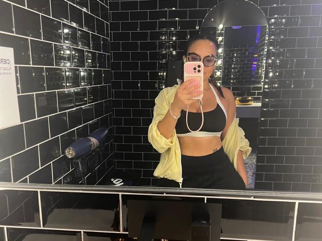 GalaMalkova with dark skin, glasses, pink phone case, black sports bra showing small tits, yellow shirt, black skirt, and earbuds, taking a mirror selfie in a black-tiled bathroom.