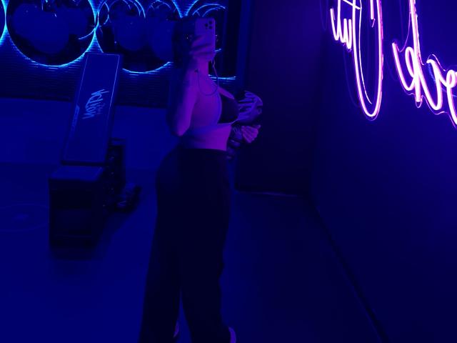 GalaMalkova stands in a dark room with blue and purple neon lights, wearing a sports bra and high-waisted pants, holding a phone, with a cute face and curvy ass.