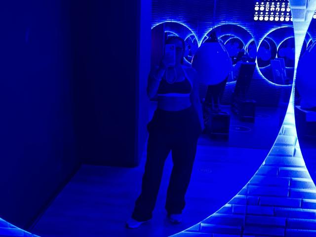 GalaMalkova with big tits in a sports bra, black pants, and white sneakers, standing in blue-lit room, arms crossed, reflected in curved mirror, glowing circular lights.