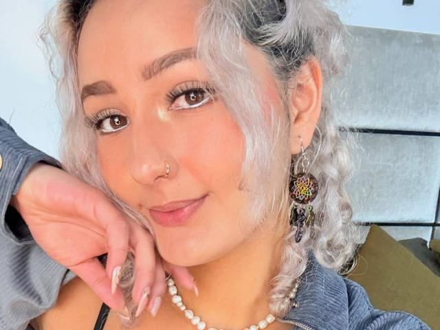 ElenaSaenz with curly gray hair, nose ring, and hoop earrings, wearing a pearl necklace, blue shirt, and holding her chin, smiling softly. Boobs not visible.