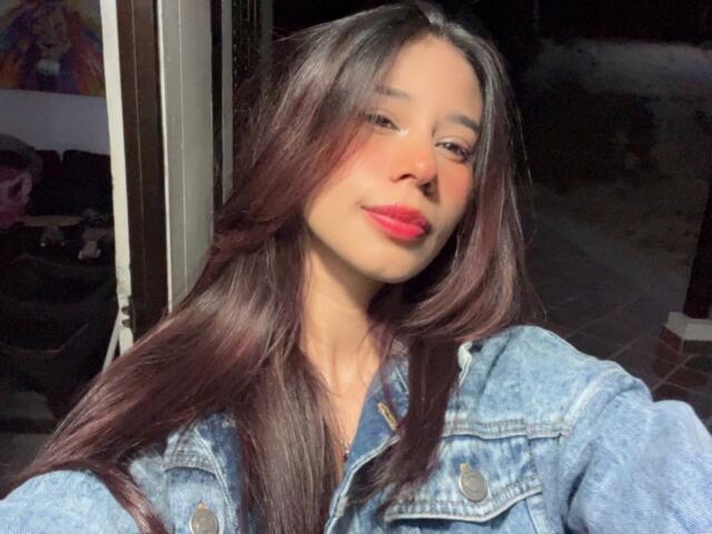 Alessandra Goldy, con lunghi capelli castano scuro, indossa una giacca di jeans, ha il rossetto rosa, sorride leggermente e la luce del sole illumina il suo viso. Le sue natiche non sono visibili..