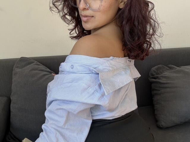 Rubyro with wavy dark red hair, glasses, nose ring, blue off-shoulder shirt, and black pants, sitting on dark gray couch, showing bare shoulder and subtle smile.