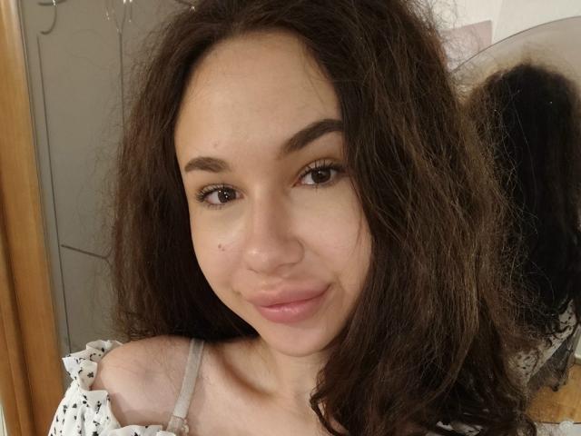 CheryLessly has wavy brown hair, brown eyes, and a small beauty mark on her right cheek. She's smiling with pink lips, wearing a white off-shoulder top.