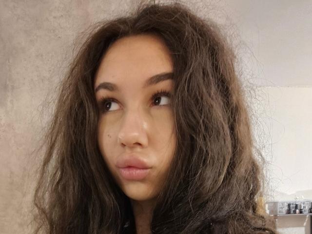CheryLessly tiene el cabello castaño despeinado, una pequeña peca en la mejilla derecha y labios rosados y gruesos. Está mirando hacia la izquierda con las cejas ligeramente levantadas..