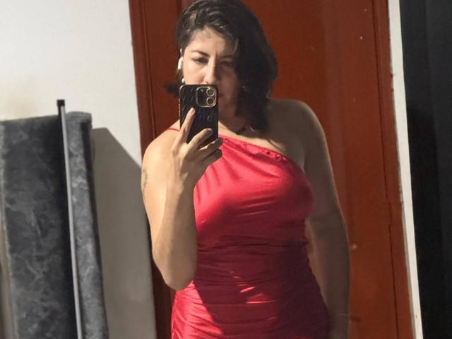 AnastasiaEvansX with medium tits, wearing a red, one-shoulder dress, holding a phone, taking a mirror selfie, brown wavy hair, standing in front of a red door.