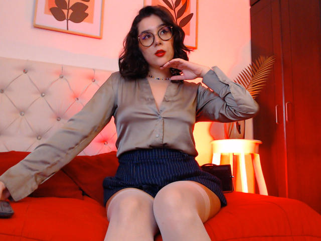 GoddessBar with glasses, red lipstick, and dark curly hair, wearing a grey button-up shirt and blue pinstripe shorts, sitting on a red bed, hand near her face.
