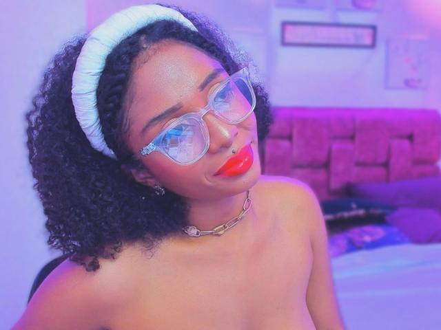 MeganWlliams with curly black hair, white headband, clear glasses, red lipstick, nose ring, chain necklace, topless, purple-blue lighting, looking at camera, confident, sexy.