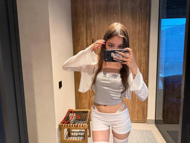 TereseGioe with long brown hair, wearing a gray crop top, white shorts, and a white cardigan, taking a mirror selfie, showing her slim waist and small tits.