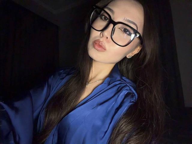 TereseGioe with long dark hair, wearing black glasses and a blue satin robe, pouting her glossy pink lips, showing off her small, cute nose and subtle earrings.