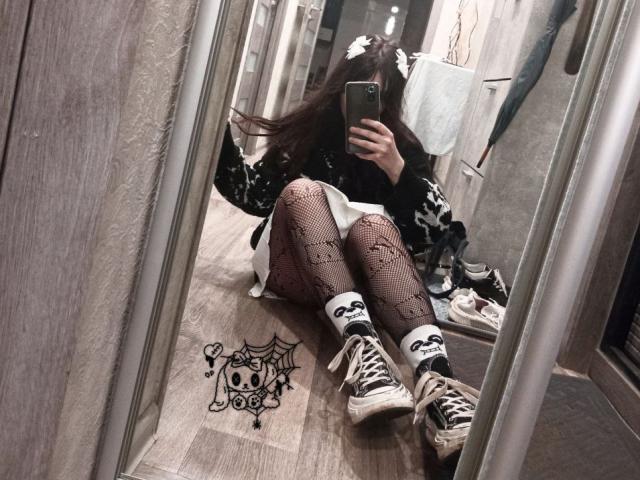 ForenCostler is sitting on the floor in front of a mirror, wearing black and white patterned tights, skull socks, and dirty black sneakers, with long brown hair.