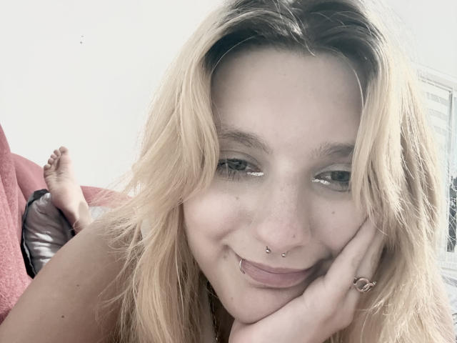 ForenCostler has blonde hair, green eyes, and a nose ring, smiling with a hand on her cheek. Her bare shoulder and feet up in the background are visible.