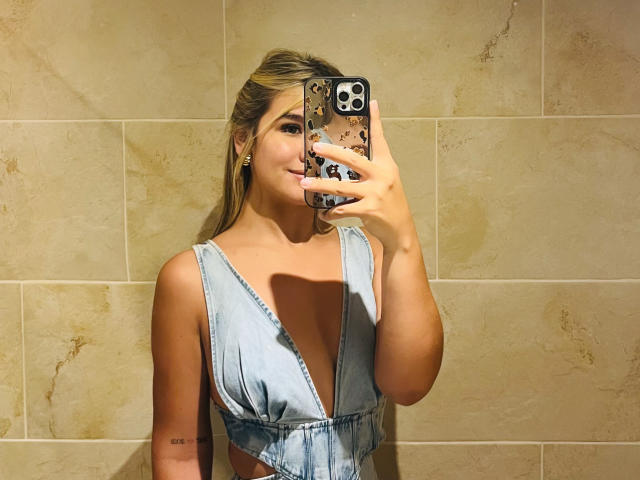 SashaPolet, met blond haar, draagt een blauwe spijkerjurk met een lage halslijn en maakt een selfie in een badkamer, waarbij haar borsten zichtbaar zijn. Ze houdt een telefoon vast met een telefoonhoesje met een patroon..