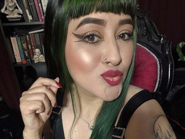 SaraEmpress, com cabelo verde, delineador preto, batom rosa, piercing no nariz e unhas vermelhas, fazendo beicinho com uma pequena gota no queixo, vestindo alças pretas..