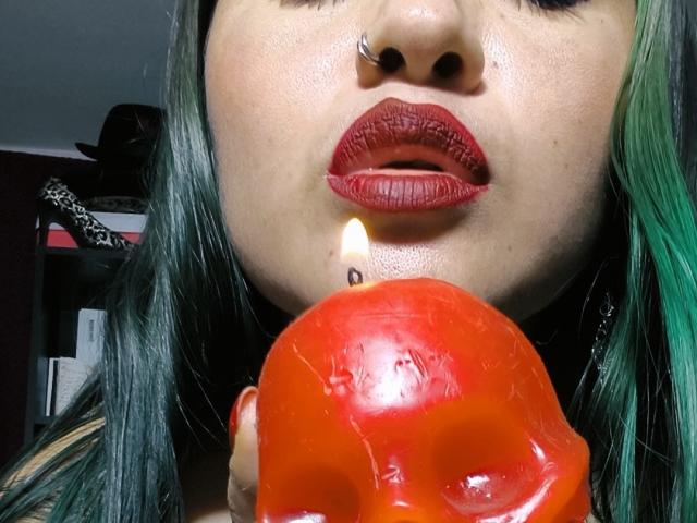 SaraEmpress with green and black hair, nose ring, and red lipstick, holding a red candle to her lips, candle flame lit.