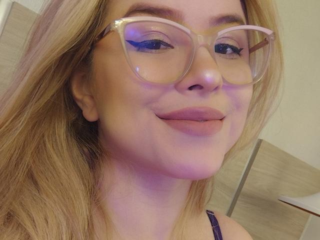 AshleyRossie with blonde hair, wearing large clear glasses, slightly smiling with her tongue out, showing her plump lips and subtle blue eyeshadow. Her tits are not visible.