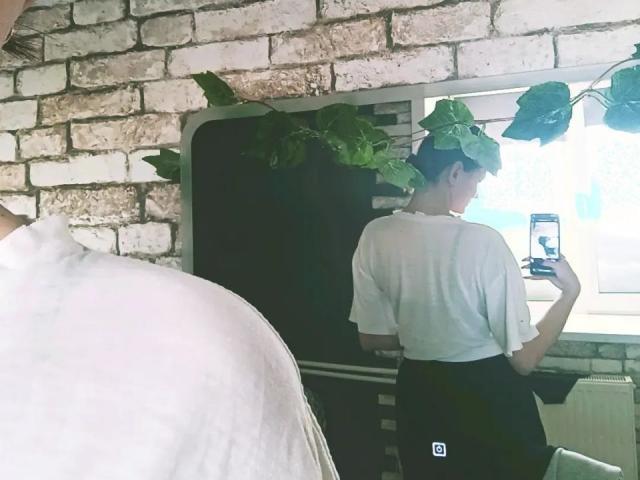LoidaGess with a white tee and black pants, taking a mirror selfie. Green leaf on head. Reflection shows phone with face. Second person's white shoulder in foreground.
