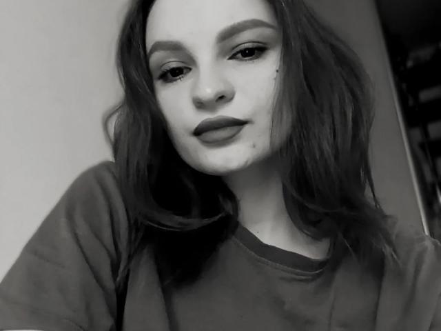 LoidaGess has a cute face, medium-length wavy dark hair, thick eyebrows, subtle makeup, dark lipstick, and a small beauty mark under her right eye. She's wearing a dark shirt.
