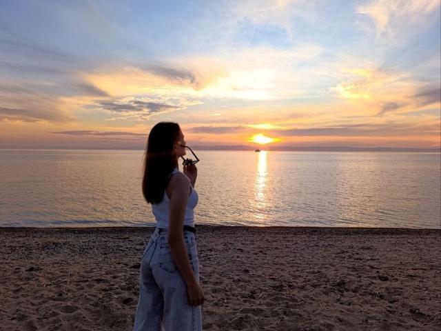 LoidaGess stands on a sandy beach, back to camera, wearing a white tank top and high-waisted blue jeans, holding sunglasses, watching a sunset over a calm ocean.